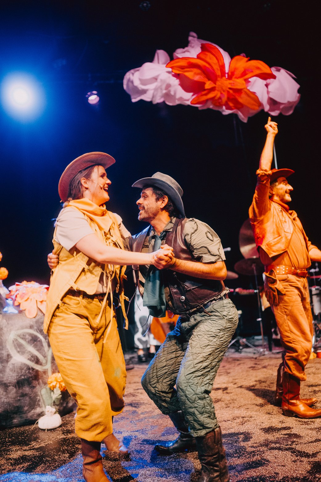 Zwei Schauspielenden tanzen einen dynamischen Paartanz im Vordergrund, sie tragen ein gelbes und ein grgünes Cowboykostüm. Über ihnen hängt eine Wolke mit großer orangener Blüte darauf. Links oben ein blaues Scheinwerferlicht.