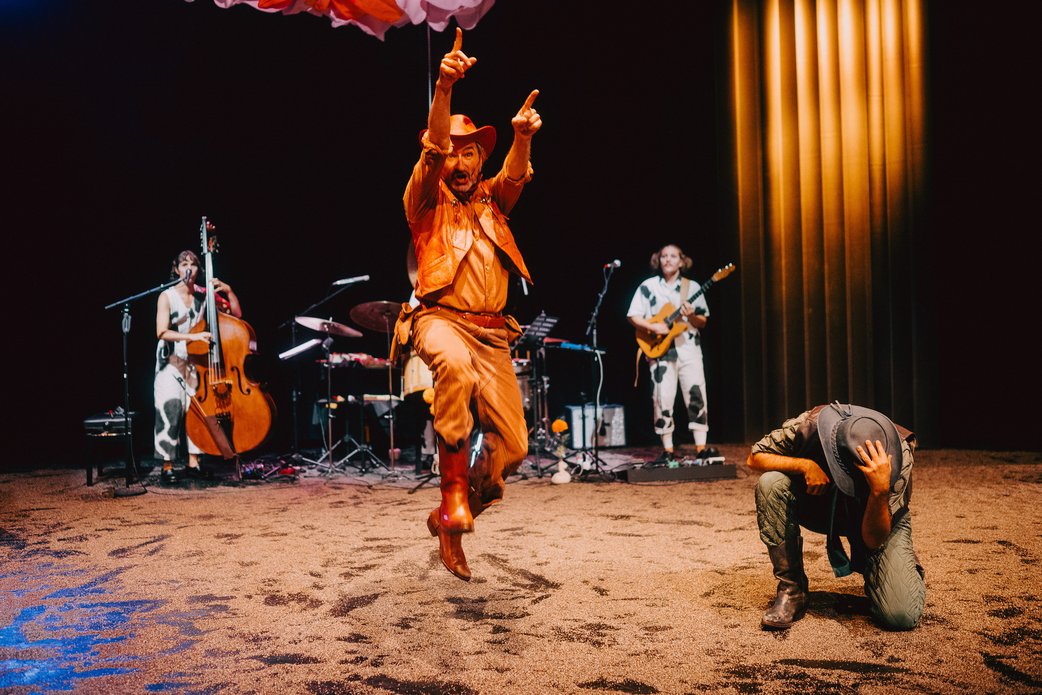 Ein Schauspieler in rotem Cowboykostüm ist frontal im Sprung zu sehen, der Bofen ist sandig, rechts ein Lamellenvorhang in hellem Licht, im Hintergrund spielt die Band. Vorn rechts bückt sich ein Schauspieler in grünem Cowboykostüm.