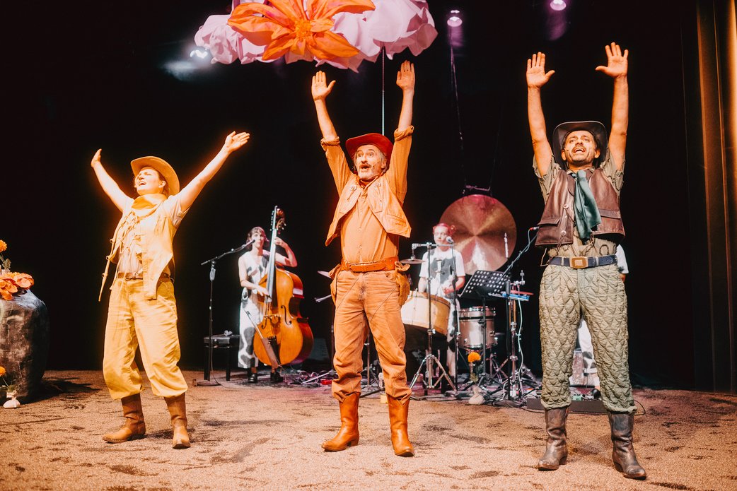 Drei Schauspielende stehen in warmen Licht nebeneinander und reißen die Arme in die Höhe, sie scheinen zu singen. Im Hintergrund die Band.