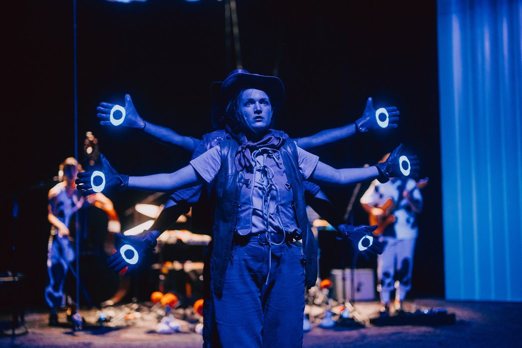 Eine Schauspielerin steht in blauem Licht (Schwarzlicht) und hat scheinbar sechs Arme in verschiedener Höhe ausgebreitet. Zwei Schauspieler stehen hinter ihr. Auf den ausgebreiteten Händen leuchten große Augen im Schwarzlicht. Im Hintergrund die Band.