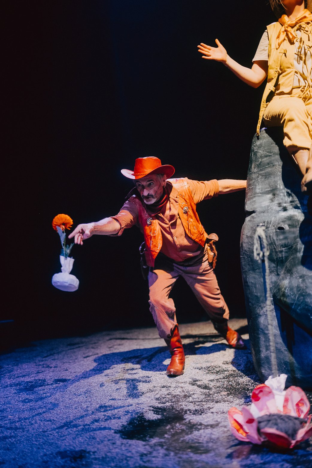 Ein Schaupieler in rotem Cowboykostüm wirft eine orangene Blum, die in einem weißen Sandsäcken steckt, wie einen Dartpfeil nach vorn. Der Boden ist sandig, der Hintergrund schwarz.