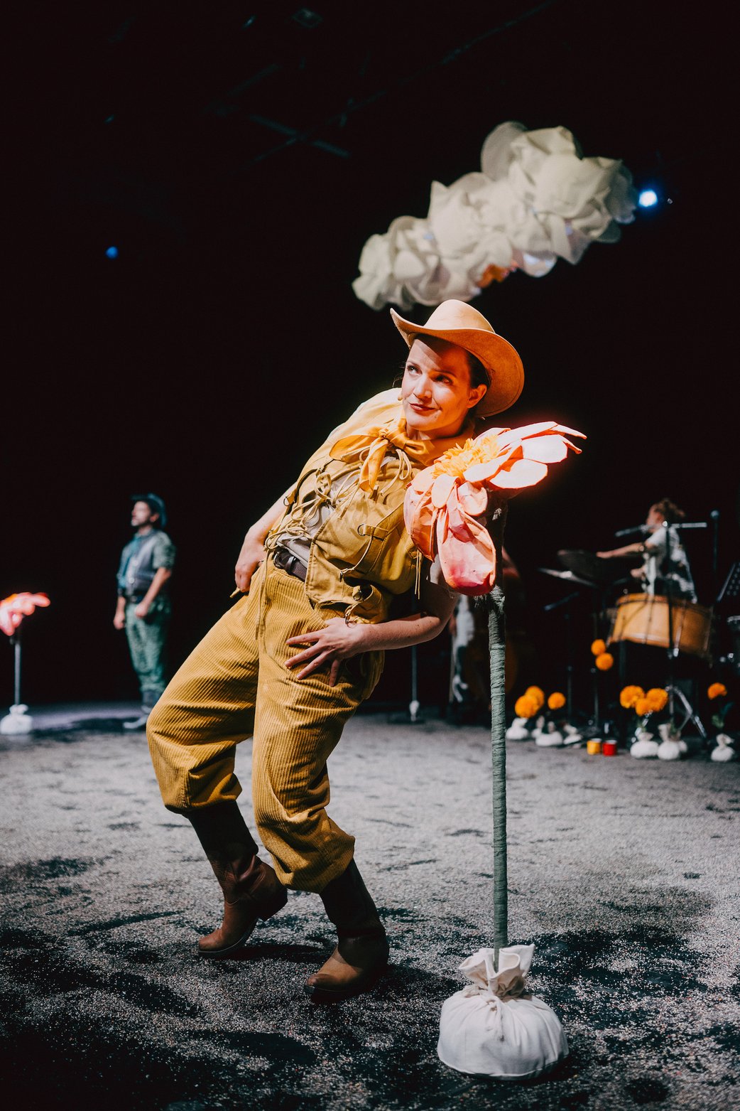 Im Vordergrund steht eine hüfthohe, orangene Blume. EIne Schauspielerin in gelbem Cowboykostüm beugt sich seitlich nach hinten zu Blüte um daran zu riechen.