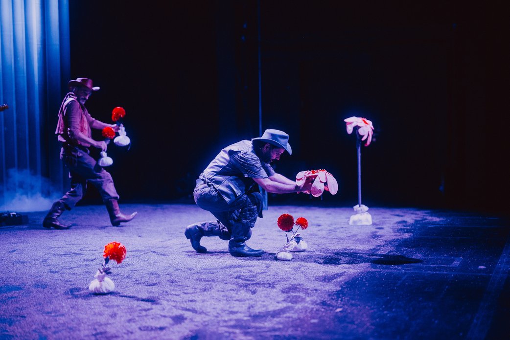 Zwei Schauspieler bringen orangene Blumen in kleinen weißen Sandsäckchen auf die Bühne. Der Boden ist sandig, die Lichtstimmung blau.