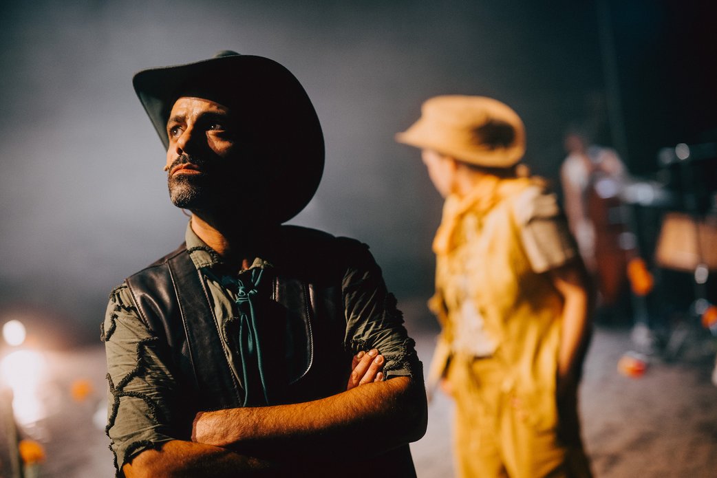 Im Vordergrund links schaut ein Schauspieler mit Cowboyhut und verschränkten Armen nach links in die Ferne. Sein Gesichtsausdruck ist emotional. Rechts wendet sich eine Schauspielerin in gelb von ihm ab, sie ist unscharf.