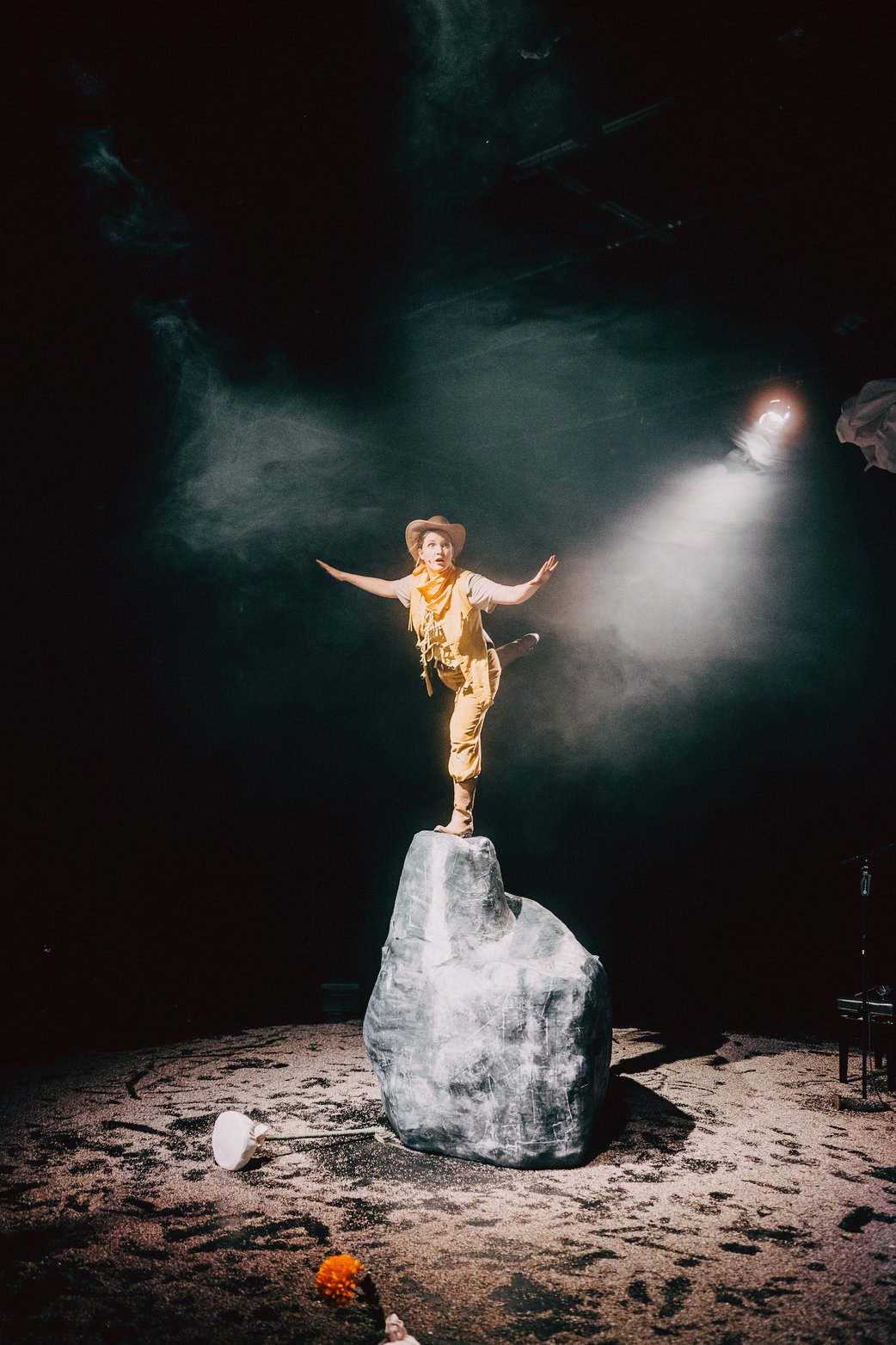 Die Schauspielerin in gelbem Cowboykostüme macht auf der Spitze eines mannshohen Felsen eine Standwaage. Scheinwerferlicht von rechts bricht sich im Nebel.